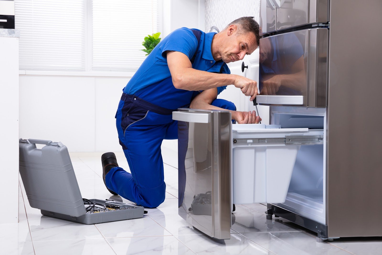 Service technician repairing an open stainless-steel refrigerator beside a tool case