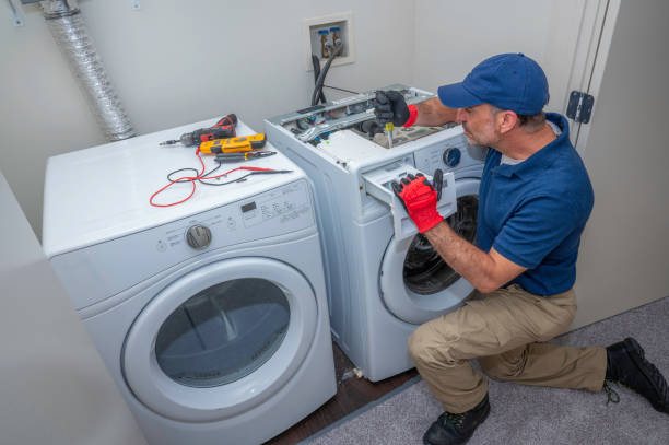 Appliance technician in blue workwear and red gloves repairing an open laundry appliance beside a dryer